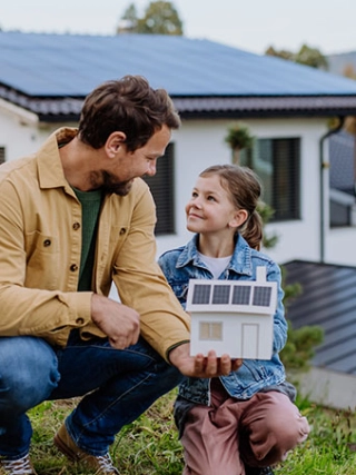 Vater mit Kind vor Ihrem Eigenheim mit einem Solardach und einem Hausmodell in der Hand.